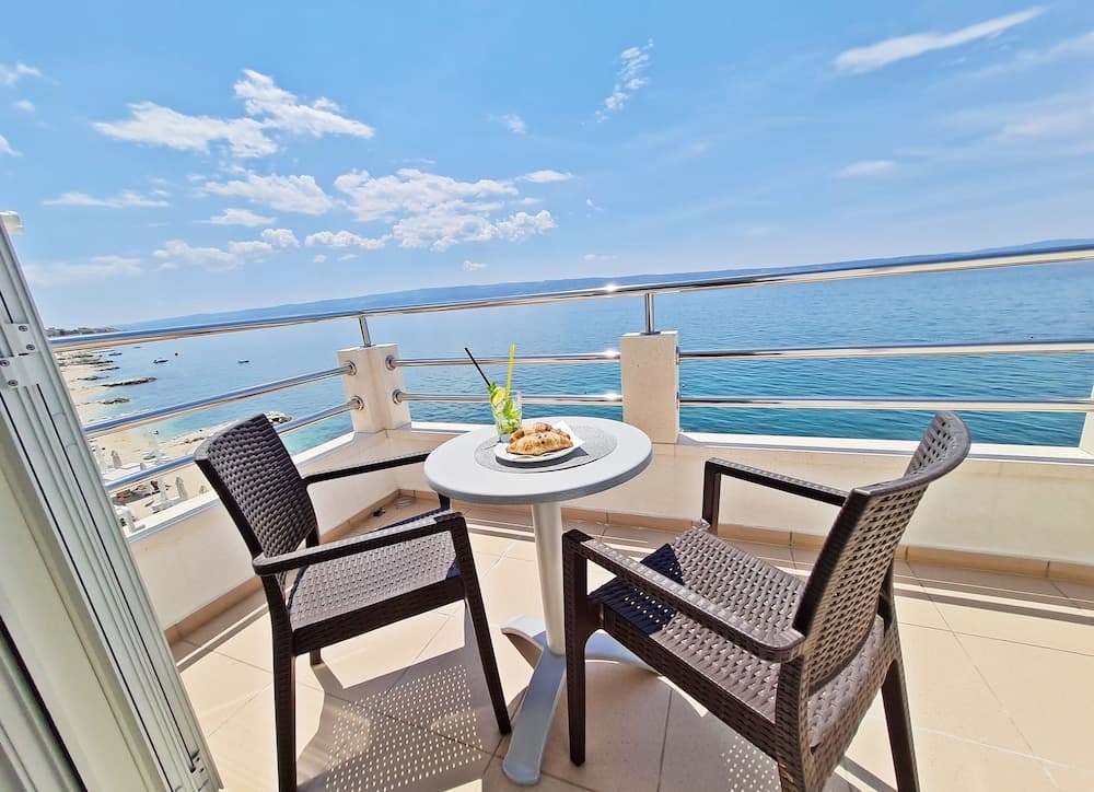 A serene seaside balcony offers a tranquil escape with wicker chairs, a small table set for breakfast, and breathtaking ocean views under a clear blue sky, perfect for a relaxing vacation getaway.