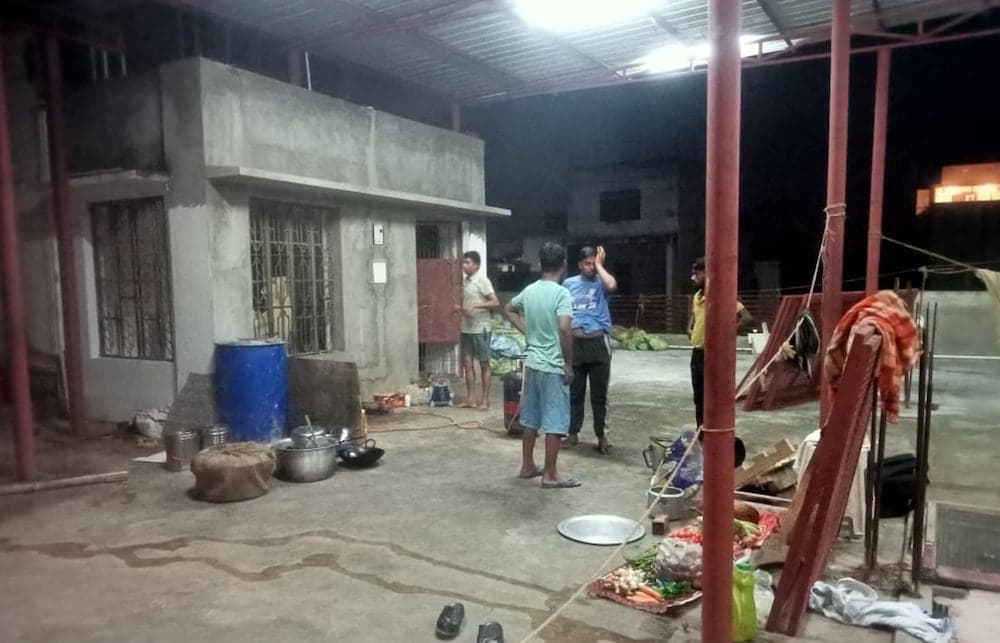 A nighttime outdoor gathering in an industrial or semi-urban area features casual interactions among people amidst scattered cooking utensils, vegetables, and laundry, ideal for exploring local culture and community life.