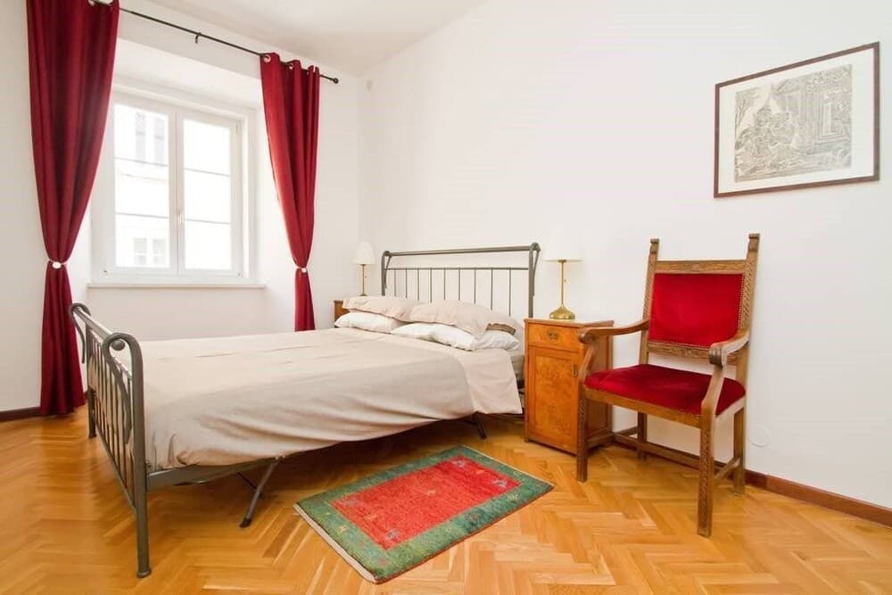 A cozy bedroom with a metal bed, red velvet chair, wooden nightstand, and vibrant rug on parquet flooring, ideal for travelers seeking a charming and comfortable stay in a quaint European apartment.