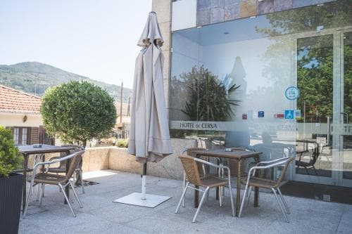 A serene outdoor seating area with tables, chairs, and a closed umbrella invites travelers to relax at a modern hotel entrance, set against a backdrop of lush green hills, ideal for leisurely stays or business retreats.