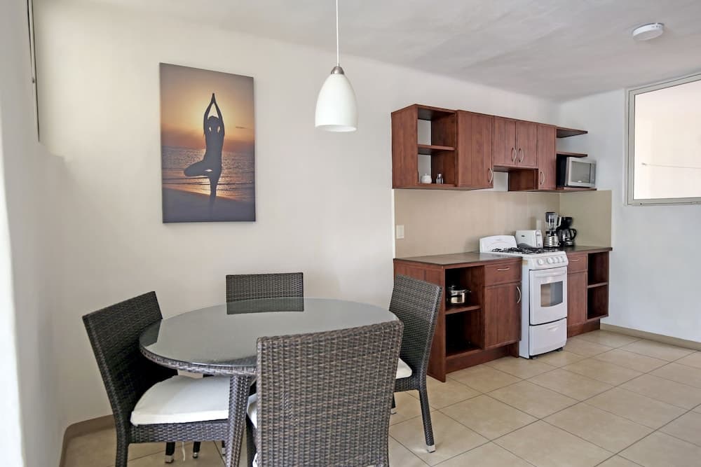 A cozy apartment kitchenette features a dining table with wicker chairs under a modern pendant light, wooden cabinetry, and a serene yoga artwork, ideal for travelers seeking a comfortable self-catering stay.