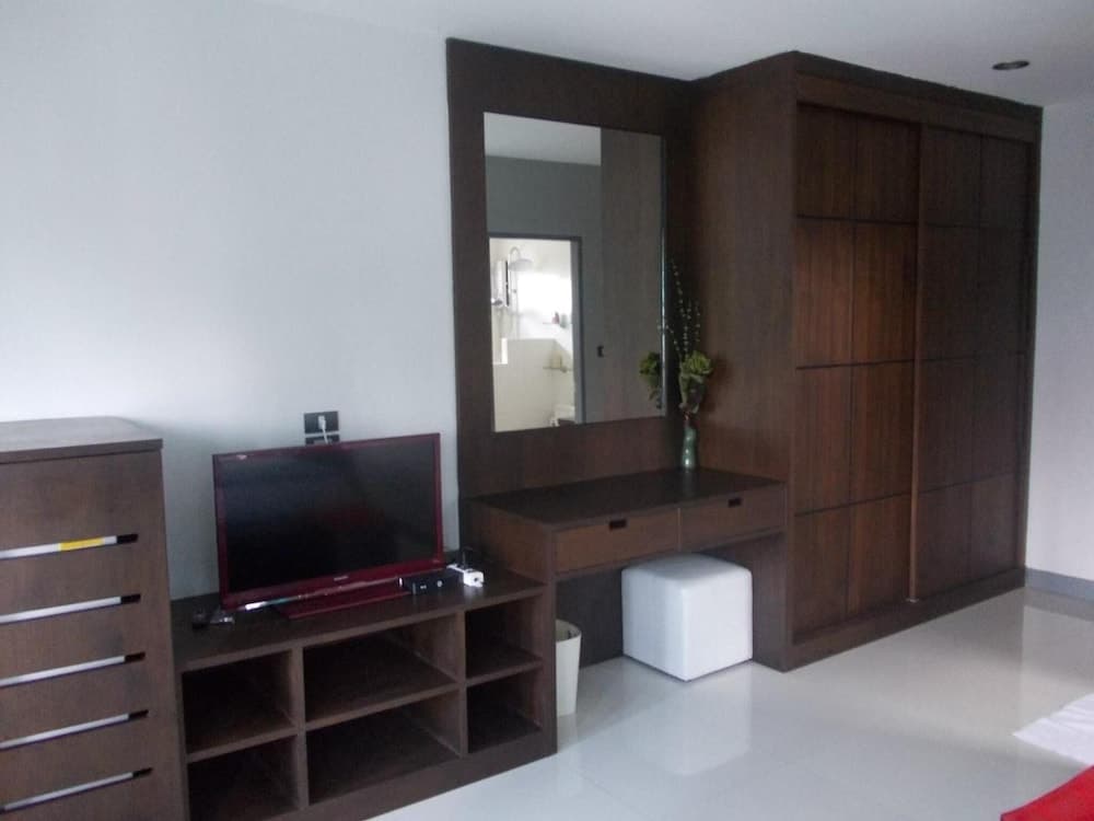 A cozy hotel room features a sleek wooden entertainment unit with a TV, a mirrored vanity with a vase of greenery, and a minimalist white ottoman, ideal for travelers seeking comfort and functionality.