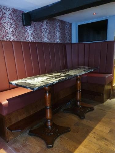 A cozy pub corner features a polished wooden table with a dark marble top set against plush red cushioned benches, under patterned wallpaper and a rustic beam, ideal for intimate dining or quiet conversations.