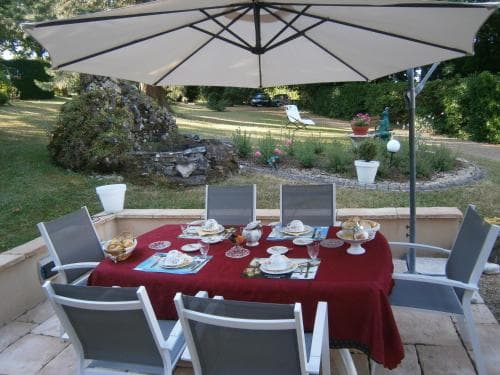 A serene outdoor dining area features a table set under a large umbrella on a wooden deck, surrounded by lush greenery and garden decor, ideal for a relaxing vacation retreat.