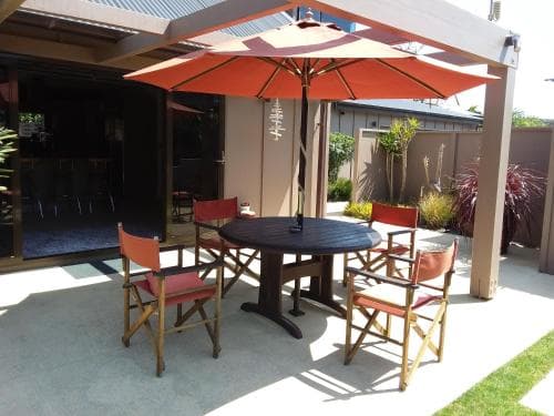 A cozy outdoor patio features a round table with red chairs under an orange umbrella, set against a modern building backdrop, ideal for relaxing or dining al fresco in a serene suburban setting.