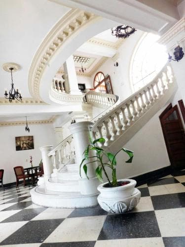 A grand, sunlit staircase with ornate balusters and a checkered floor creates an elegant atmosphere in a historic building, ideal for travelers seeking luxurious accommodations or event venues.