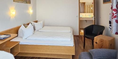 A cozy hotel room features a neatly made double bed with white linens, wooden furniture, warm wall sconces, and an adjacent bathroom, ideal for travelers seeking comfort and convenience.