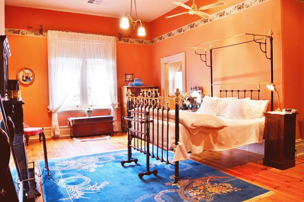 A cozy, warmly-lit bedroom with orange walls, a vintage iron bed, wooden floors, and a blue patterned rug creates an inviting atmosphere ideal for a relaxing getaway in a historic inn.