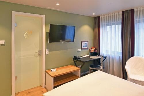 A cozy hotel room features a neatly made bed, a desk with a chair, a flat-screen TV, and a glass door leading to a bathroom, ideal for travelers seeking comfort and convenience.