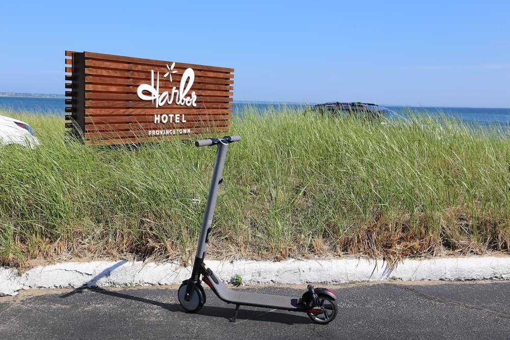 A serene coastal scene features an electric scooter parked on a paved path near lush grass, with a wooden "Harbor Hotel" sign and the ocean in the background, ideal for eco-friendly beachside exploration.