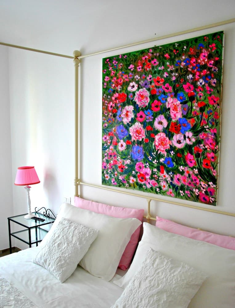 A cozy bedroom features a vibrant floral painting above a neatly made bed with white and pink pillows, complemented by a charming pink lamp on a glass side table, ideal for travelers seeking a serene and artistic retreat.