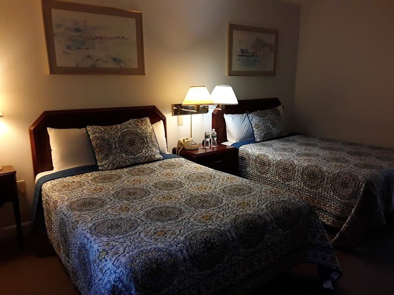 A cozy hotel room at the Castle Manor Inn in Gloucester, MA, features two patterned beds under warm lighting, framed artwork, and a bedside table with a phone, ideal for travelers seeking comfort and relaxation.