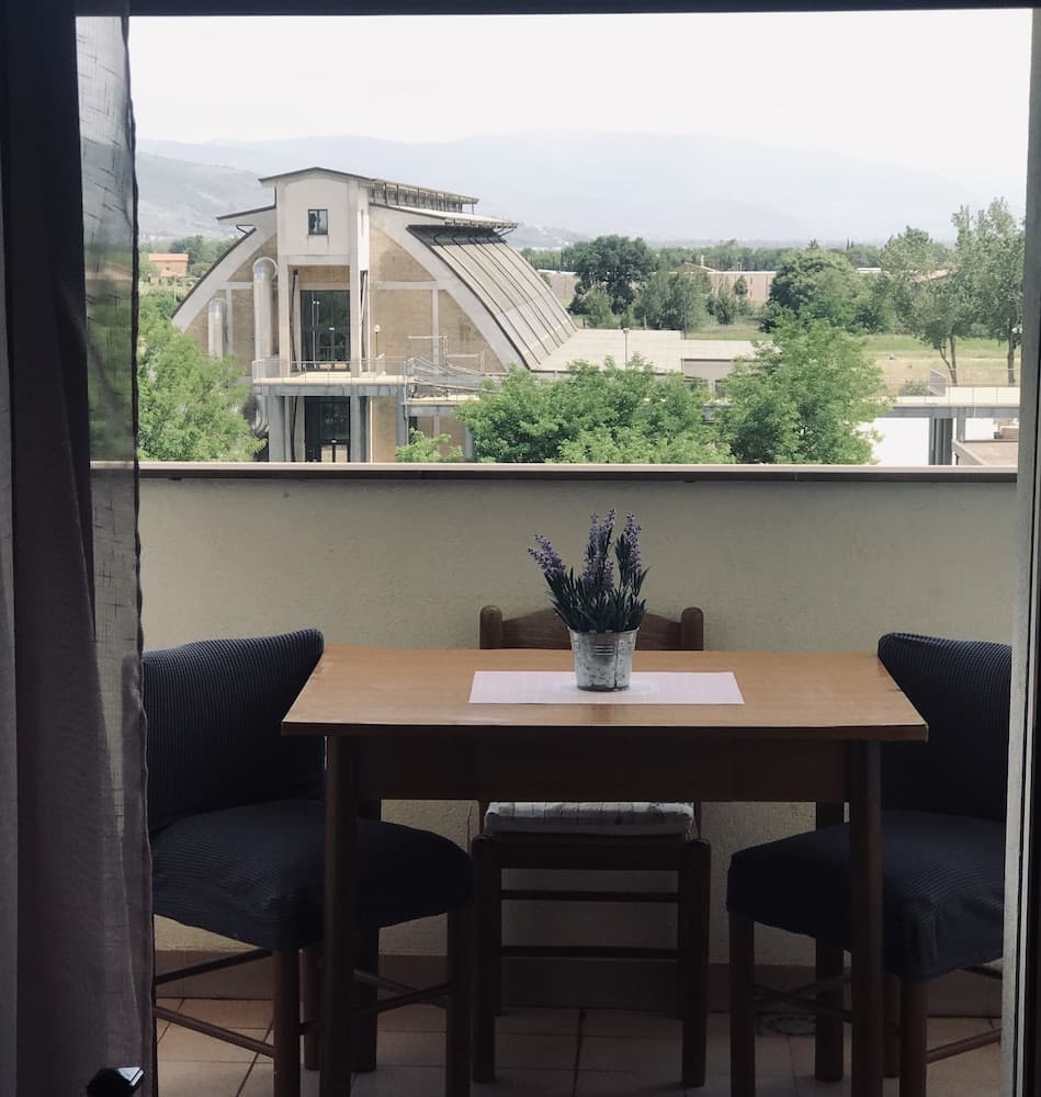 A serene balcony view features a cozy table set with lavender in a vase, overlooking lush greenery and a modern building against a backdrop of distant mountains, ideal for travelers seeking a tranquil retreat.