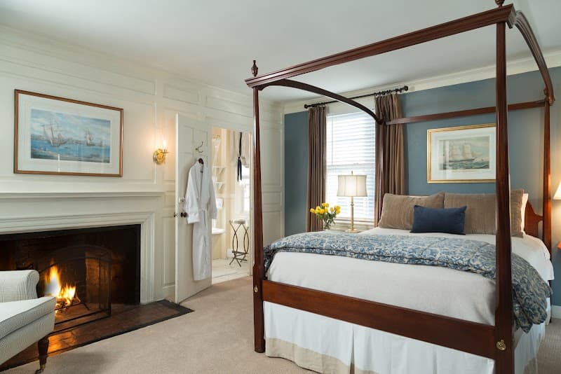 A cozy inn room features a four-poster bed, a warm fireplace, nautical artwork, and a robe hanging by the door, ideal for travelers seeking a charming and relaxing stay in Marblehead, MA.