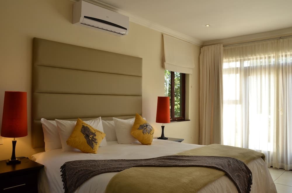 A cozy hotel bedroom features a neatly made bed with yellow pillows, red lamps on side tables, sheer curtains letting in soft light, and an air conditioner, ideal for travelers seeking comfort and relaxation.
