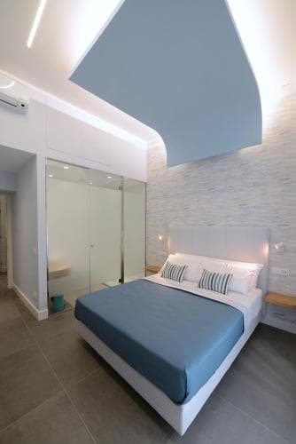 A modern bedroom with sleek white furniture, a blue bedspread, and an adjacent glass-enclosed shower creates a serene and stylish retreat ideal for travelers seeking contemporary accommodations.