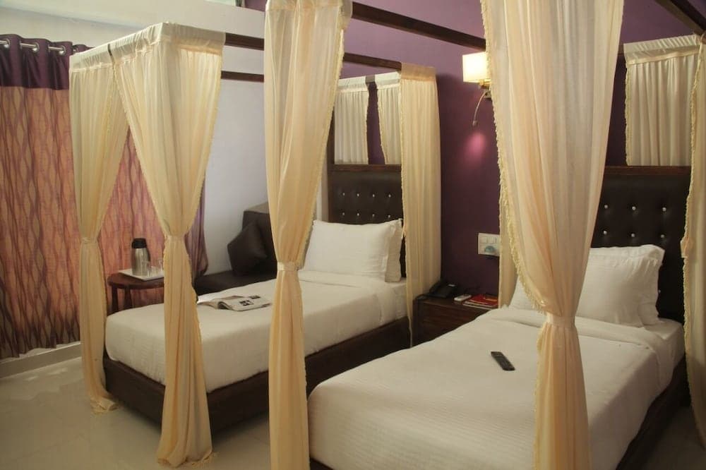 A cozy hotel room features two elegant canopy beds with cream drapes, purple walls, and modern furnishings, ideal for travelers seeking comfort and style.