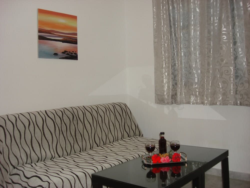A cozy living room with a striped sofa, a glass table adorned with wine glasses and flowers, a serene sunset painting on the wall, and patterned curtains, ideal for travelers seeking a relaxing and intimate accommodation setting.