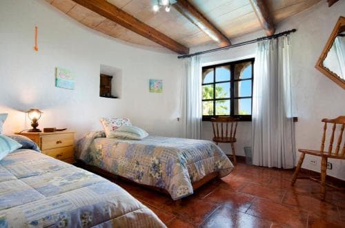 A cozy bedroom with terracotta floors, wooden beams, and a sunny window view of palm trees, ideal for a relaxing retreat in a rustic villa.