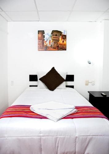 A cozy hotel room features a neatly made bed adorned with colorful textiles, a framed cityscape painting on the wall, and minimalist decor, ideal for travelers seeking comfort and simplicity.