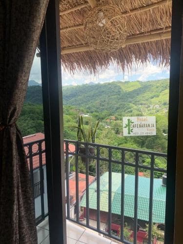A serene balcony view of lush green mountains framed by a thatched roof and vibrant rooftops, ideal for travelers seeking a tranquil retreat in a tropical setting.