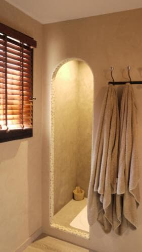 A serene bathroom with a rustic arched doorway leading to a small sink area, featuring soft beige walls, wooden blinds, and neatly hung towels, ideal for travelers seeking a tranquil spa-like retreat.