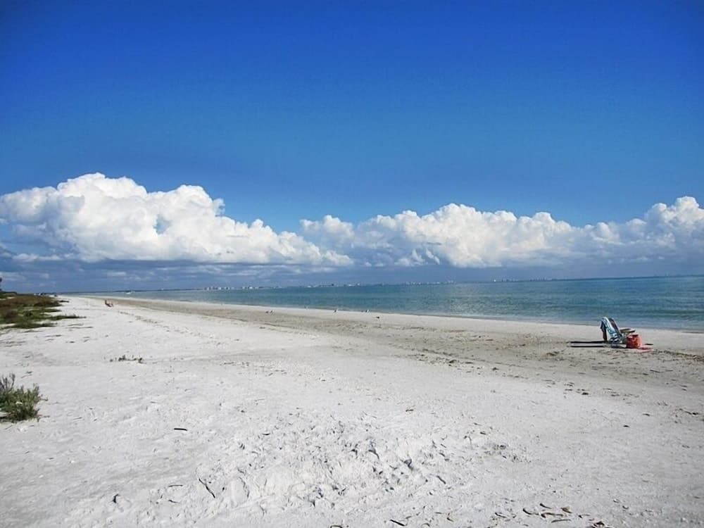 A serene beach scene with pristine white sand, calm turquoise waters, scattered lounge chairs, and fluffy clouds under a vibrant blue sky, ideal for relaxation and coastal getaways.