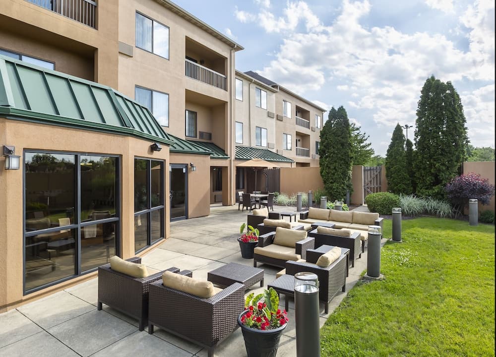 A serene hotel patio features comfortable wicker seating, lush greenery, and potted flowers under a partly cloudy sky, ideal for travelers seeking relaxation and outdoor leisure.