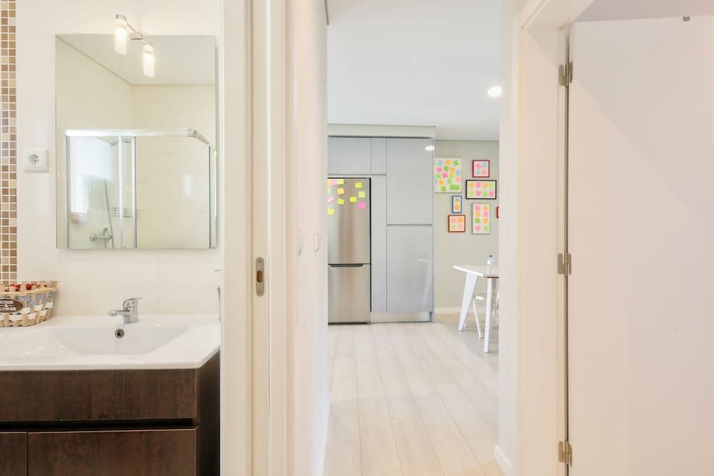 A cozy apartment hallway leads from a modern bathroom with a sleek sink and mirror to a bright kitchen with stainless steel appliances and colorful wall decorations, ideal for travelers seeking a comfortable and stylish short-term rental.