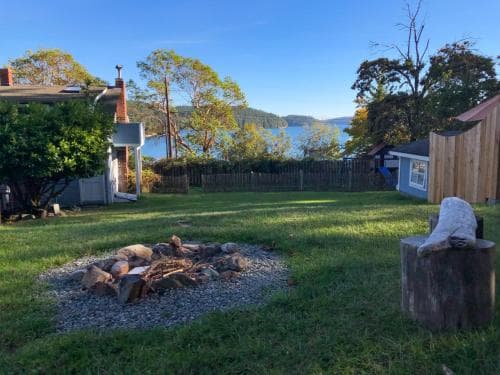 A serene lakeside backyard features a cozy fire pit surrounded by lush greenery, a rustic wooden fence, and a stone sculpture, ideal for tranquil retreats and nature-inspired relaxation.