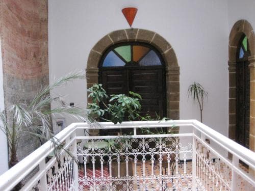 A serene indoor courtyard features arched stained-glass windows, lush potted plants, and ornate white railings, ideal for travelers seeking a tranquil retreat in a historic building.