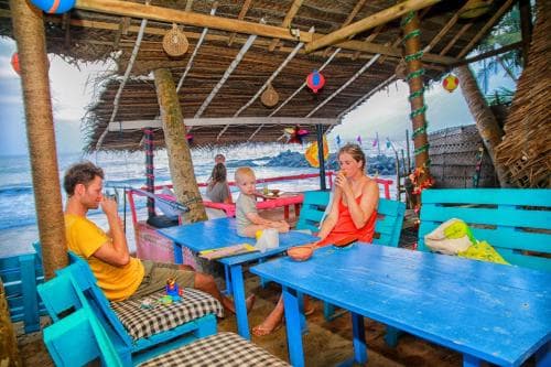 A vibrant beachside café with blue tables and chairs under a thatched roof offers a relaxed seaside dining experience, perfect for travelers seeking a casual tropical getaway.