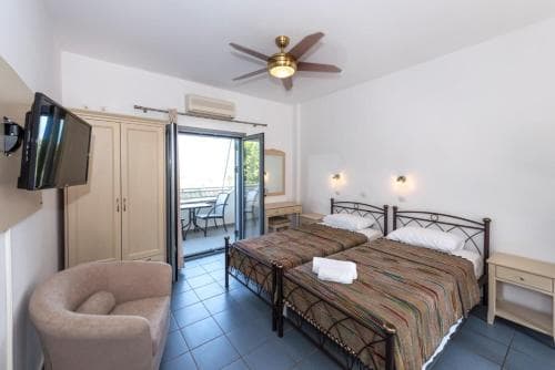 A cozy hotel room with tiled floors, a double bed adorned with striped bedding, a balcony view, and modern amenities like a mounted TV, ideal for travelers seeking comfort and relaxation.