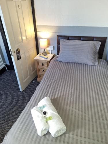 A cozy hotel room features a neatly made bed with striped bedding, a rolled towel on top, a wooden bedside table with a lamp, and a welcoming ambiance ideal for travelers seeking comfort and relaxation.