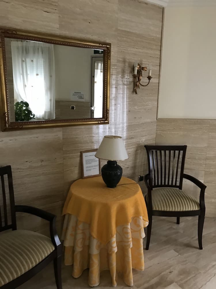 A cozy hotel lobby corner features marble walls, elegant wooden chairs with striped cushions, a yellow tablecloth-covered table adorned with a lamp and vase, a gold-framed mirror reflecting sheer curtains, and a warm sconce light, ideal for travelers seeking a serene waiting area.