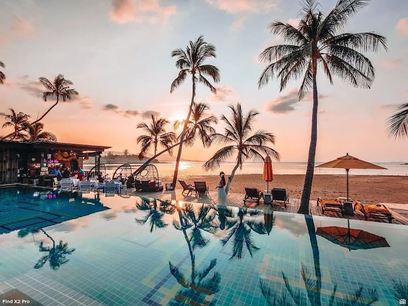 A serene sunset at a luxurious beachfront resort features palm trees, an infinity pool reflecting the sky, lounge chairs, and a vibrant bar area, ideal for travelers seeking relaxation and tropical ambiance in Thailand.