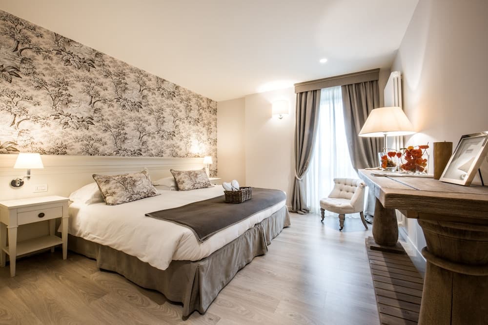 A cozy hotel room features a neatly made bed against a botanical wallpaper backdrop, soft lighting from bedside lamps, a plush armchair by the window, and a rustic wooden desk, ideal for travelers seeking comfort and relaxation.