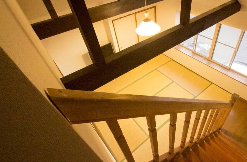 A cozy traditional room with tatami flooring, wooden beams, and a warm pendant light viewed from a staircase, ideal for travelers seeking authentic cultural accommodations.