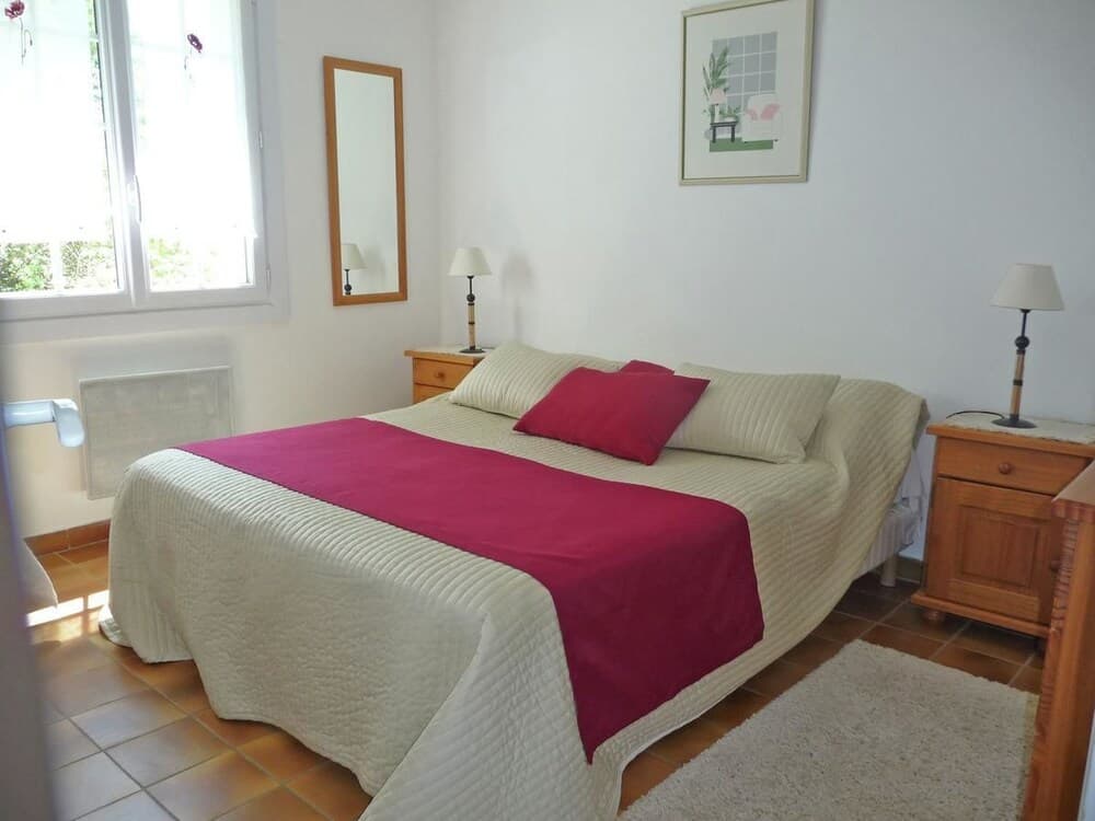 A cozy bedroom with a neatly made bed adorned in cream and magenta linens, wooden nightstands with lamps, a mirror, and a window letting in natural light, ideal for travelers seeking a serene and comfortable stay.