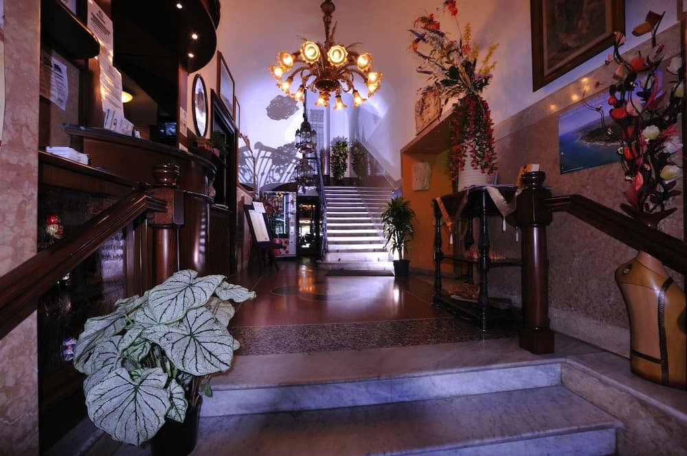 A warmly lit hotel lobby features elegant marble stairs, lush greenery, artistic decor, and a glowing chandelier, creating an inviting atmosphere ideal for travelers seeking luxury and comfort.