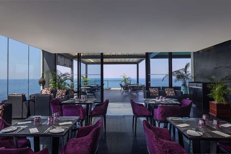 A luxurious dining area at the Radisson Hotel in Colombo offers a serene coastal view through expansive glass windows, featuring elegant purple chairs and tables set for an intimate meal, ideal for travelers seeking a refined dining experience with ocean vistas.