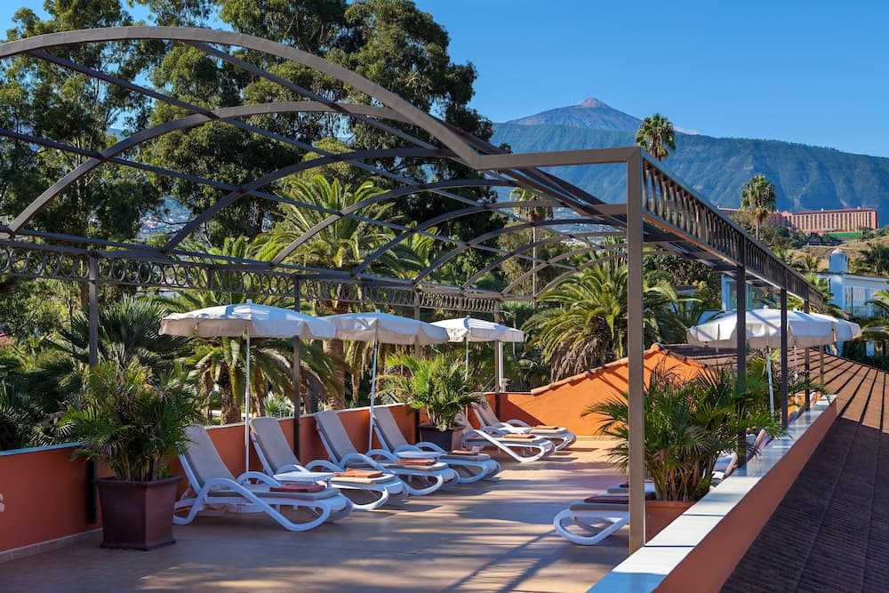 A serene rooftop terrace lined with lounge chairs under umbrellas, surrounded by lush palm trees and a stunning mountain backdrop, offers an ideal spot for relaxation during a tropical getaway.