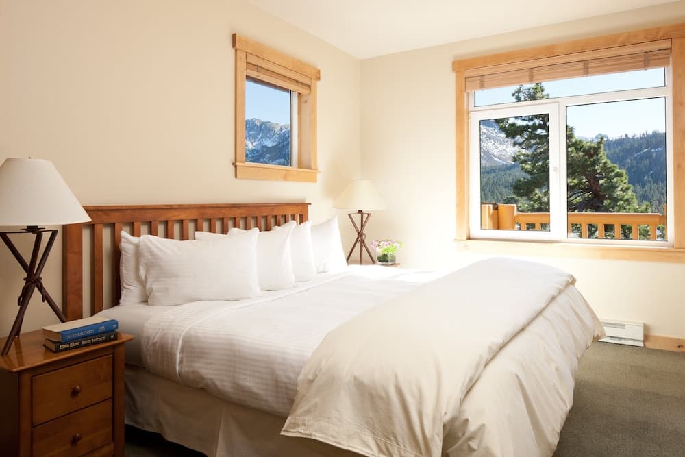 A cozy hotel bedroom with a neatly made bed, wooden furnishings, and large windows offering breathtaking views of snow-capped mountains and lush greenery, ideal for travelers seeking relaxation in a serene mountain retreat.