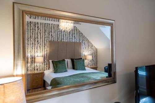 A cozy hotel room features a large mirror reflecting a neatly made bed with green bedding, wooden bedside tables, and a birch-patterned wall, creating an inviting atmosphere ideal for travelers seeking comfort and relaxation.