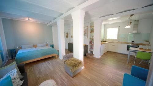 A cozy open-plan apartment features a bedroom with a teal bedspread, a modern kitchen with white cabinets, and vibrant blue chairs, creating a welcoming and stylish space ideal for a relaxing getaway.
