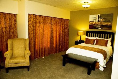 A cozy hotel room features warm-toned curtains, a neatly made bed with a bench at its foot, a comfortable armchair, and soft lighting, creating an inviting atmosphere ideal for travelers seeking relaxation.