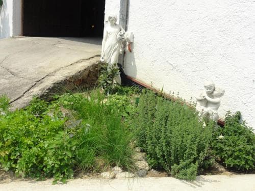 A serene garden corner beside a white building features lush greenery, stone statues of a standing figure and a child, evoking a peaceful atmosphere ideal for spiritual retreats or quiet contemplation.