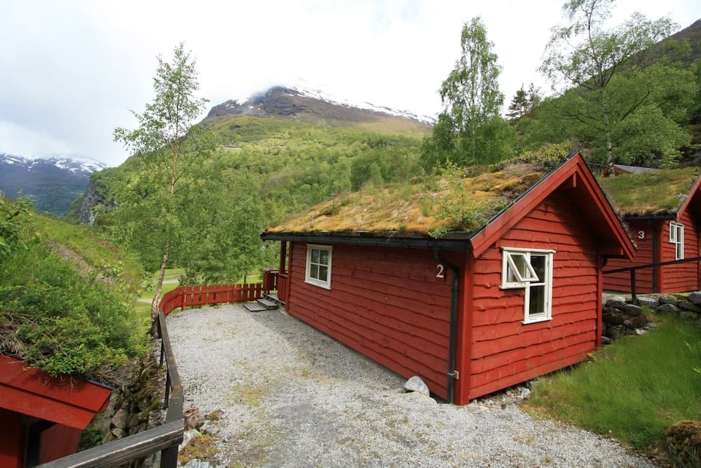 A serene red cabin with a moss-covered roof nestles amidst lush greenery and towering mountains, offering a tranquil retreat for nature lovers seeking a cozy getaway.