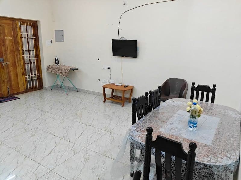 A cozy dining area in a villa features a round table with black chairs, a wall-mounted TV, and a marble floor, ideal for travelers seeking comfort in Puducherry's MG Road Area.