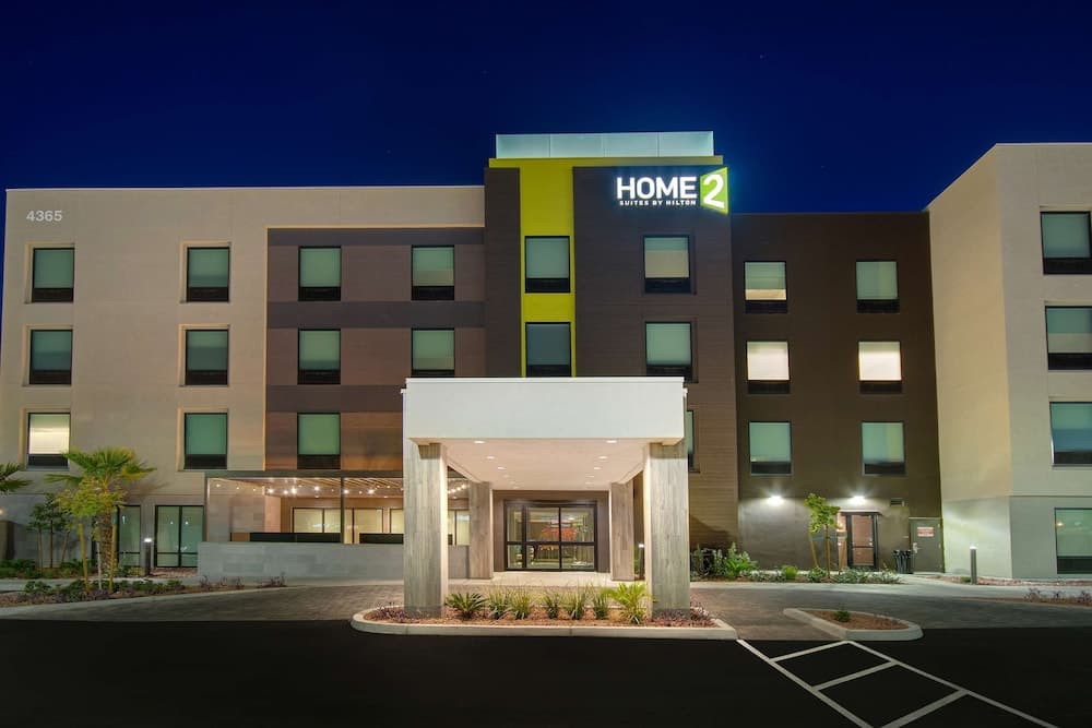 A modern Home2 Suites by Hilton hotel stands illuminated at night, featuring a welcoming entrance, landscaped grounds, and a calm atmosphere ideal for travelers seeking comfortable accommodations.
